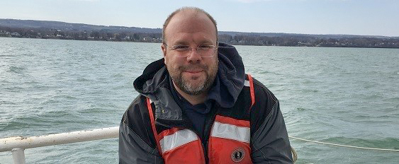 Man wearing a life jacket and jacket standing on a boat with a calm body of water and distant shoreline in the background. The sky is overcast, and the man is smiling slightly, looking toward the camera.